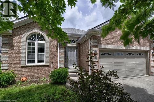 Brick exterior featuring a two-car garage with arched window inserts, an arched entry window with louvered shutters, a gray front door, and mature landscaping